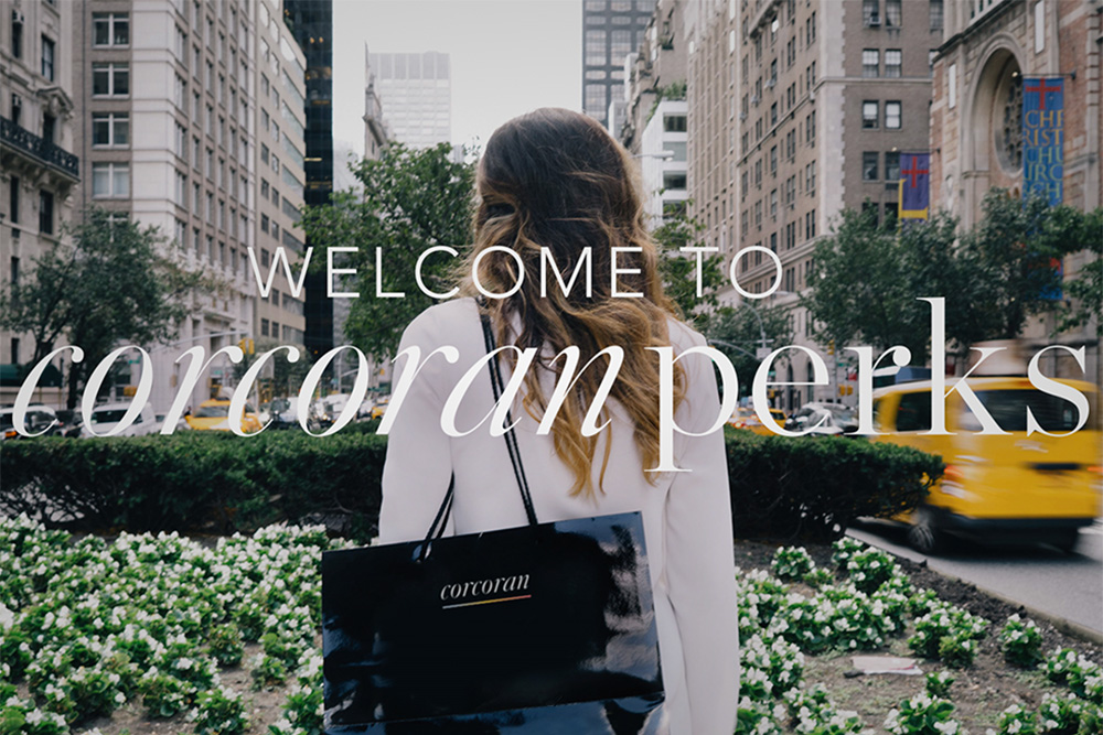 Test says Welcome to CorcoranPerks over a woman carrying a bag standing in the middle divider of large street in a large city