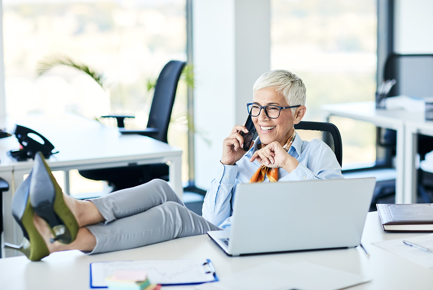 Mature woman sitting at her desk with her feet up talking on a cell phone and smiling.